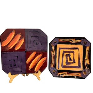 May include: Two decorative ceramic plates with abstract designs. One plate is divided into four sections, with orange brushstrokes on a burgundy and purple background. The other plate has a yellow and purple spiral design on a black background.