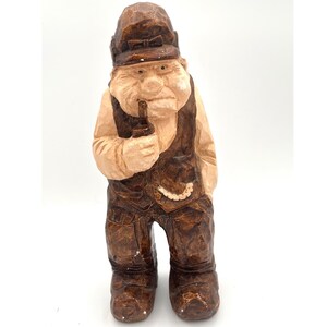 May include: A hand-carved wooden figurine of a man wearing a hat and vest, holding a small bottle. The figure is painted in shades of brown and tan, with visible wood grain. The figurine has a detailed, rustic appearance.