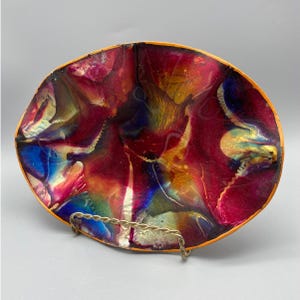 Könnte beinhalten: Eine ovale dekorative Schale mit einem lebendigen, abstrakten Design. Die Schale weist eine Mischung aus Farben auf, darunter Rot, Blau, Gelb und Orange, mit einem glänzenden Finish. Sie wird von einem Metallständer getragen und zeigt ihre künstlerische Anziehungskraft.