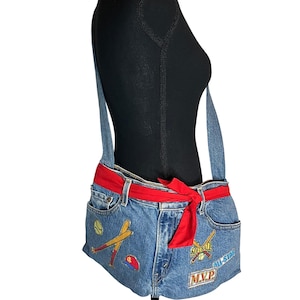 May include: A denim crossbody bag with a red fabric belt. The bag features baseball and softball patches, including the words "SOFTBALL", "ALL-STAR", and "MVP". The bag has a denim shoulder strap and two front pockets.
