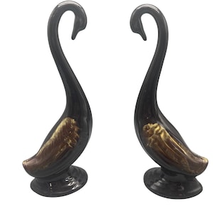 Puede incluir: Dos figuras de cisnes negros con detalles dorados en el cuerpo. Los cisnes tienen cuellos largos y curvos y están sobre una base redonda. Las figuras son de material brillante, probablemente decorativas.