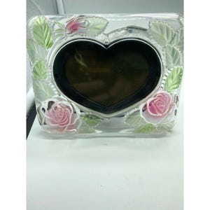 May include: Clear glass picture frame with a heart-shaped photo area. The frame is decorated with pink roses and green leaves. The frame is square and has a silver border around the heart-shaped photo area.