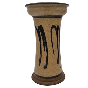 Puede incluir: Un jarrón de cerámica beige con la parte superior e inferior ensanchadas. El jarrón presenta líneas verticales negras y curvas y un borde y base marrón oscuro. El jarrón tiene una superficie texturizada y forma cilíndrica.