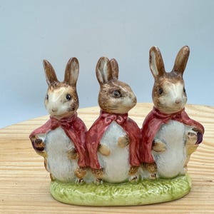May include: A ceramic figurine featuring three brown and white rabbits standing side by side on a green base. Each rabbit wears a red cape tied at the neck. The rabbits have pointed ears, dark eyes, and white bellies, crafted in a glossy finish.
