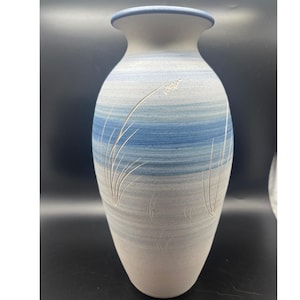 Może przedstawiać: Wysoki, ceramiczny wazon z szeroką podstawą i rozszerzonym brzegiem. Wazon posiada poziome pasy w kolorze jasnoniebieskim i szarym, z wytrawionymi wzorami kwiatowymi. Obrzeże jest ciemnoniebieskie. Dekoracyjny element wystroju domu.