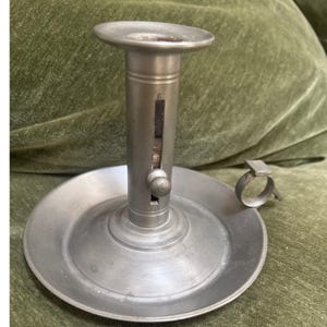 May include: A silver-toned metal chamberstick candle holder featuring a wide circular base, a central cylindrical shaft with a vertical adjustment slot, a small sliding knob, and a side-mounted finger loop handle for carrying.