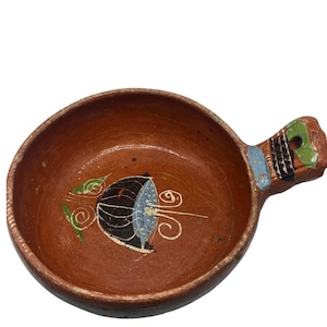 May include: A small, round, terracotta-colored bowl with a handle. The bowl features a painted design of a black object with blue and white accents, along with green leaves and white swirls. The handle has blue, black, and green painted details.