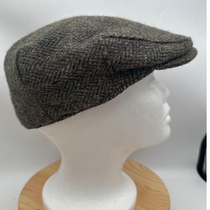 Puede incluir: Una gorra plana con un patrón de espiga texturizado en tonos marrón oscuro, gris y sutiles motas de color óxido. La gorra tiene una corona estructurada y una visera corta y rígida, confeccionada en un tejido con apariencia de lana.