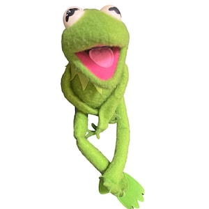 以下が含まれることがあります： 鮮やかな緑色のボディ、黒い瞳の大きな白い目、ピンクの口が特徴のカーミット・ザ・フロッグの人形。人形は大きく口を開けており、腕を組んだポーズをとっています。カエルは黄色の襟と緑色の足ひれを持っています。