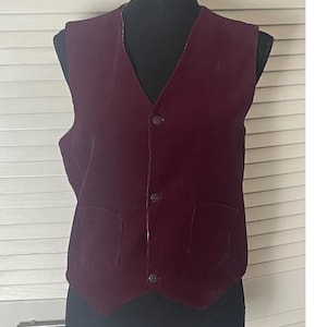 May include: A deep burgundy corduroy waistcoat featuring a V-neckline, a front button fastening with three matching buttons, and two patch pockets at the lower front. The garment is sleeveless and has a pointed hemline at the centre front.