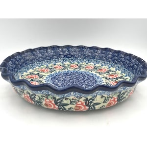 May include: A round ceramic pie dish with a scalloped edge, featuring a dark blue rim and a central pattern of red floral motifs surrounded by intricate blue and white decorative scrollwork and small leaf details across the entire surface.