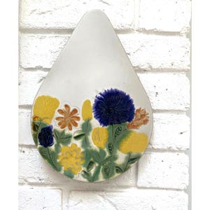 Può includere: Un'applique da parete in ceramica a forma di goccia con un motivo floreale. I fiori sono gialli, arancioni e blu, con foglie verdi. Lo sfondo è bianco, su un muro di mattoni bianchi.