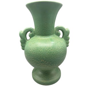 Può includere: Un vaso di ceramica verde con una texture maculata. Il vaso ha un corpo bulboso, un bordo svasato e due manici curvi. Poggia su una base a piedistallo e il design è simmetrico e decorativo.