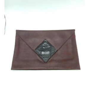 May include: A brown leather envelope clutch bag with a black diamond-shaped detail. The clutch has a flap closure and a simple design. The leather appears smooth, and the overall aesthetic is minimalist and stylish.