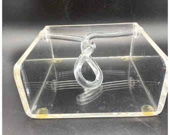 Op de afbeelding: Een heldere, zeshoekige acryl doos met een gedraaide handgreep. De doos heeft een transparant ontwerp, waardoor de inhoud zichtbaar is. Kleine, ronde, goudkleurige voetjes zijn zichtbaar aan de onderkant van de doos. De handgreep is een doorlopende lus.