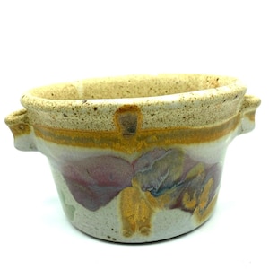 May include: A handmade ceramic bowl with two side handles. The bowl features a speckled tan rim, a yellow and white band, and a body with purple, yellow, and blue abstract designs. The bowl is suitable for serving or display.