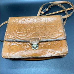 May include: A tan leather crossbody bag with a floral embossed design. The bag features a flap closure with a metal clasp and a long, adjustable strap. The stitching is white, and the leather has a slightly distressed appearance.
