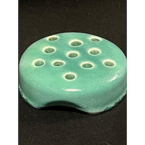 May include: A round, seafoam green ceramic flower frog with multiple holes for arranging stems. The frog has a slightly textured surface and a curved base, designed to sit in a vase. The holes are evenly spaced across the top.
