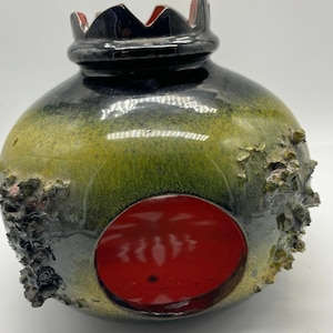 May include: A ceramic pomegranate-shaped vase with a glossy finish. The vase features a dark green and black exterior with a large red circular opening. The top has a crown-like design, and the surface has textured, rough patches.