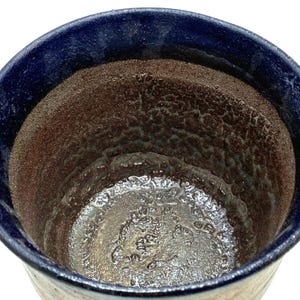 Puede incluir: Primer plano de una taza de cerámica con un borde azul oscuro y un interior texturizado. El interior de la taza tiene un aspecto moteado marrón y plateado, con una superficie rugosa e irregular. El exterior de la taza es de color beige claro.