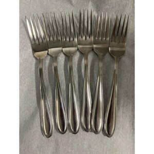 May include: A collection of six stainless steel forks. Each fork features a long, slender handle with a rounded base and a tined head. The forks are arranged in a line, with their tines facing upwards. The forks are on a light grey surface.