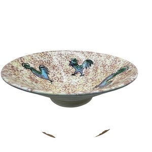 Könnte beinhalten: Eine flache Keramikschale mit einem Sockelfuß, die eine cremefarbene Oberfläche mit braunen Sprenkeln aufweist. Das Innere ist mit drei handbemalten Hähnen in Blau- und Grüntönen verziert, die in einem kreisförmigen Muster angeordnet sind.