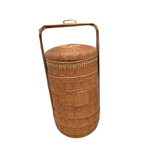 May include: A woven brown basket with a handle. The basket is cylindrical with multiple stacked compartments and a domed lid. The handle is arched and attached to the sides of the basket. The basket is made of natural fibers.