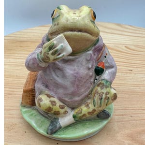 Peut inclure: Une figurine de grenouille en céramique portant une veste violet clair, assise sur une base verte. La grenouille a une tête jaune pâle, des yeux sombres et des pattes tachetées. Elle tient un petit objet rectangulaire blanc et un détail orange.