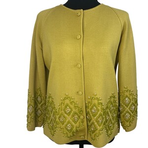 Puede incluir: Un cárdigan de color chartreuse con mangas largas y cuello redondo. El suéter con botones presenta intrincados detalles bordados en tonos verdes y blancos a lo largo de los puños y el dobladillo inferior. El bordado tiene un patrón geométrico.