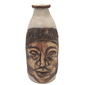 May include: A handmade ceramic vase with a face motif. The vase has a light beige top, a dark brown band, and a detailed face with closed eyes and a moustache. The colour scheme includes shades of brown and beige, creating a unique decorative piece.
