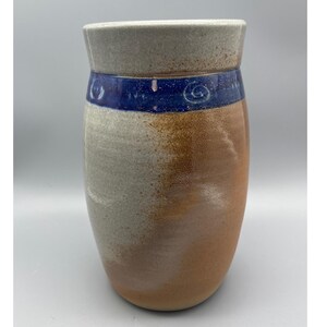 May include: A handmade ceramic vase with a unique design. The vase features a light gray rim, a dark blue band with spiral designs, and a two-tone body in shades of brown and gray. The vase has a rounded shape and a textured surface.