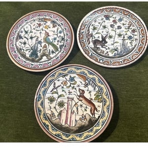 Könnte beinhalten: Drei runde Keramikteller mit handbemalten Motiven. Ein Teller zeigt einen Vogel, ein anderer einen Hasen und der dritte einen Hund. Jeder Teller ist mit komplexen Blumenmustern, stilisierten architektonischen Elementen und dekorativen Rändern versehen.