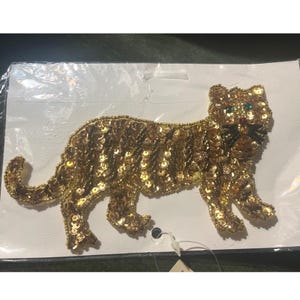 以下が含まれることがあります： 金色のスパンコールタイガーブローチ。虎は歩行姿勢で、体は金色のスパンコールで覆われています。目は緑色です。ブローチは白い背景にあります。