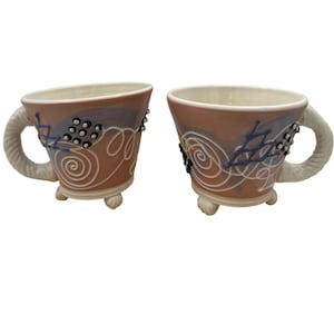 Puede incluir: Dos tazas de cerámica con base marrón claro, con patrones de espirales blancas, detalles azules y una cuadrícula de puntos en relieve. Cada taza tiene un asa texturizada y retorcida, y se apoya sobre tres pequeños pies redondeados.