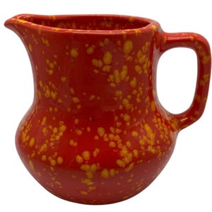 Op de afbeelding: Een rode keramische kan met een geel gespikkeld patroon. De kan heeft een gebogen lichaam, een tuit en een handvat. Het ontwerp is een levendig, opvallend stuk voor het serveren van drankjes of als decoratief item.