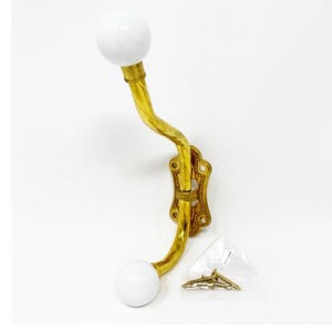 May include: A wall-mounted coat hook featuring a curved, polished brass arm with a textured, twisted design. The hook has two smooth, white spherical ceramic knobs at each end and a decorative brass mounting plate with four screw holes.