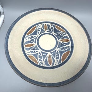 May include: A circular ceramic plate with a cream-coloured centre and a blue rim. The centre has a blue and brown leaf-like pattern around a white circle. The outer edge is cream-coloured with a dark blue border.