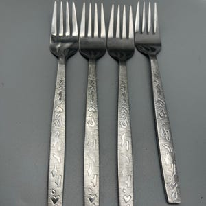 May include: Four stainless steel forks with a textured design on the handles. The handles feature a pattern of shapes and hearts. The forks are arranged in a row against a gray background.