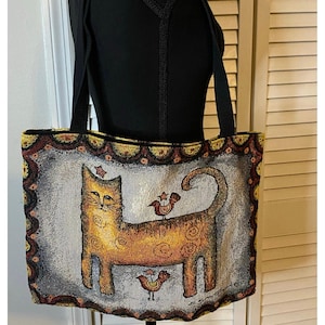 May include: Tote bag with a tapestry design featuring a stylized orange cat with two birds on its back. The bag has black straps and a decorative border with gold and burgundy accents. The cat has a star above its head.