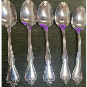 Pode incluir: Cinco colheres de prata com um desenho floral na base do cabo. As colheres têm uma superfície refletora e estão dispostas em fila. As colheres provavelmente fazem parte de um conjunto de talheres.