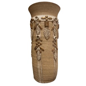 May include: A tall, beige ceramic vase with a wide opening and decorative relief patterns. The vase features leaf and floral designs in a lighter beige and gold colour, adding a touch of elegance to its rustic appearance. The vase is approximately 30 cm tall.