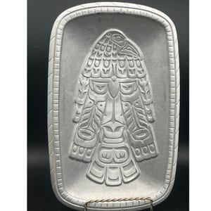 May include: A rectangular, silver-coloured decorative tray with a raised, detailed relief of a stylized bird or totem design. The tray has a textured surface and a beaded border.
