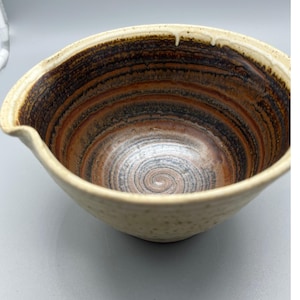 Puede incluir: Un cuenco de cerámica hecho a mano con un exterior color crema y un interior marrón oscuro y rojizo. El interior presenta un diseño en espiral. El cuenco tiene un pico vertedor.