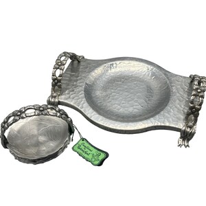 May include: Two-piece silver-toned serving set. The larger piece is a tray with handles adorned with floral designs. The smaller piece is a round dish with a floral handle. Both pieces have a textured surface.