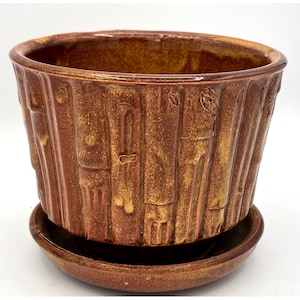 Könnte beinhalten: Ein brauner Keramik-Blumentopf mit passender Untertasse. Der Topf hat ein strukturiertes, vertikales Bambus-ähnliches Design mit glänzendem Finish. Die Farbe ist eine Mischung aus Braun und Gold, wodurch eine warme, erdige Ästhetik entsteht.
