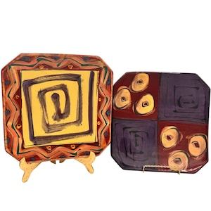 May include: Two decorative octagonal plates with abstract designs. One plate features a yellow center with a brown spiral and a patterned border. The other plate has a maroon background with purple squares and gold circular designs.