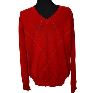 May include: A red V-neck sweater with a diamond pattern. The sweater has long sleeves and a ribbed hem and cuffs. The diamond pattern is made up of thin black and gold lines.