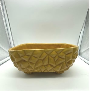 Pode incluir: Um vaso de cerâmica amarelo mostarda com um exterior geométrico e texturizado. O vaso retangular assenta em três pequenos pés. O interior é liso, contrastando com o exterior áspero. Uma peça decorativa.