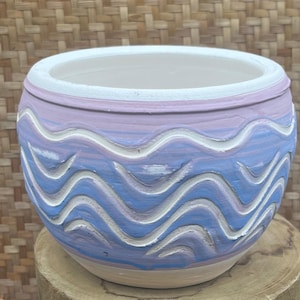 Puede incluir: Una maceta redonda de cerámica con un diseño ondulado. La maceta está pintada en tonos de azul y lavanda, con patrones de olas blancas talladas. El borde es blanco y el interior es de color beige claro. La maceta está sobre una superficie de madera.