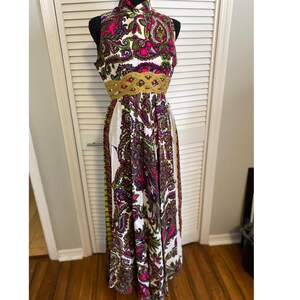 May include: A sleeveless, floor-length dress with a high neckline. The dress features a vibrant paisley pattern in pink, purple, green, and white. A gold-colored embellished belt cinches the waist. The dress is displayed on a mannequin.
